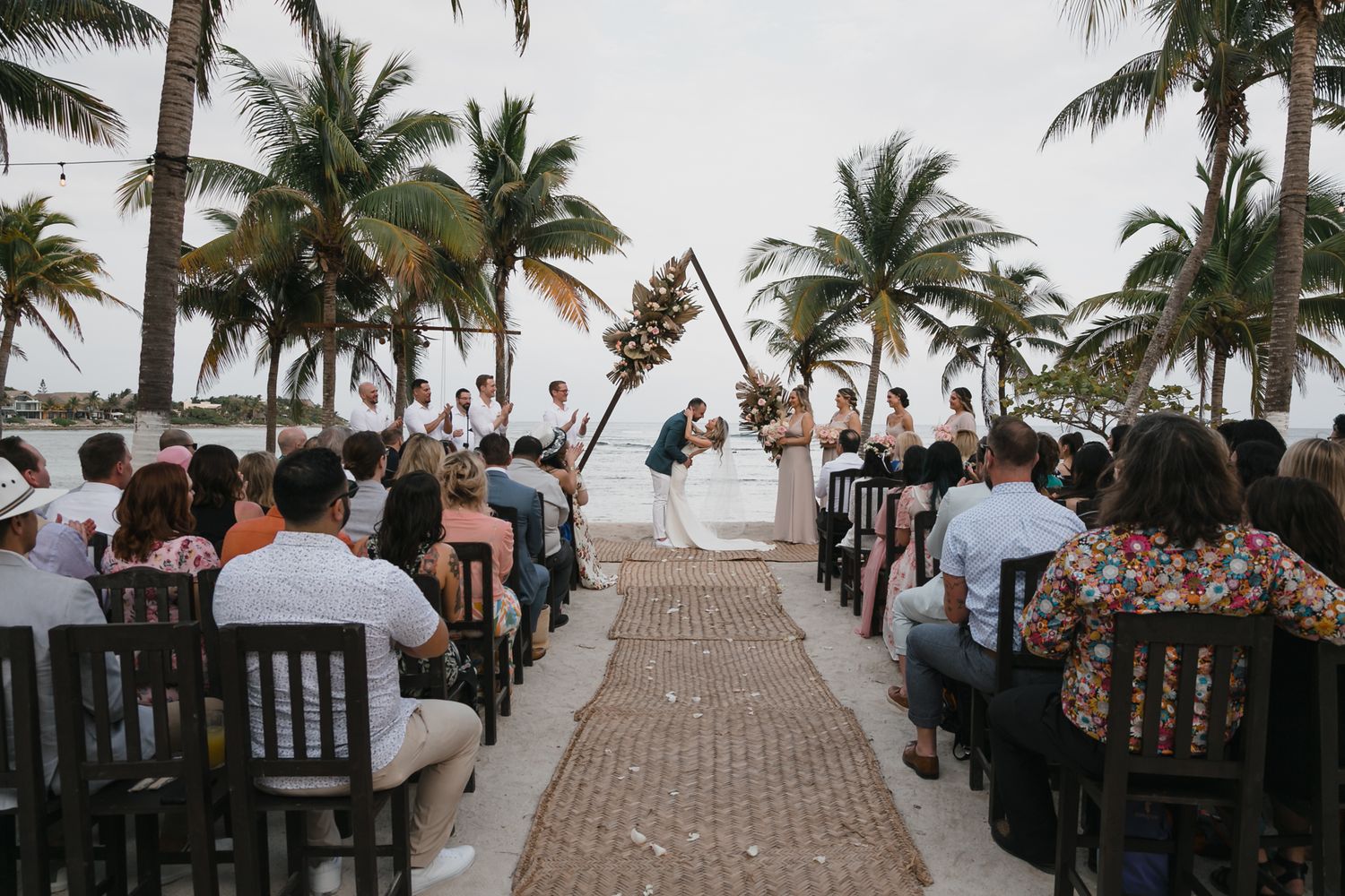Tropical beach wedding ceremony with guests seated along a rustic aisle leading to a triangular floral arch.