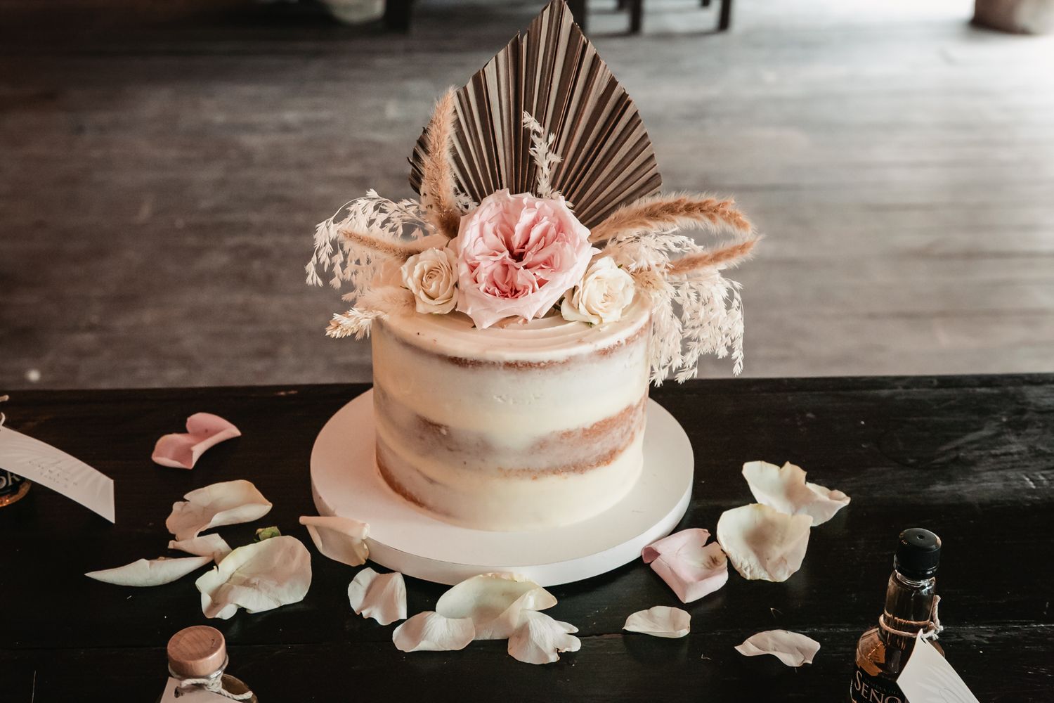 Elegant semi-naked wedding cake topped with pink roses and dried palm leaf on decorated table.