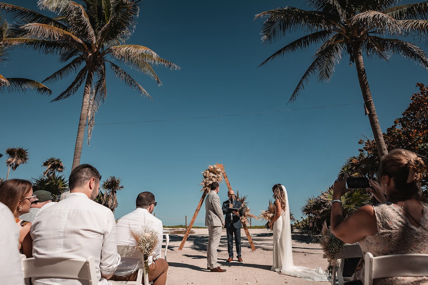 Palm trees line a beachfront wedding ceremony with guests in white and neutral colors.