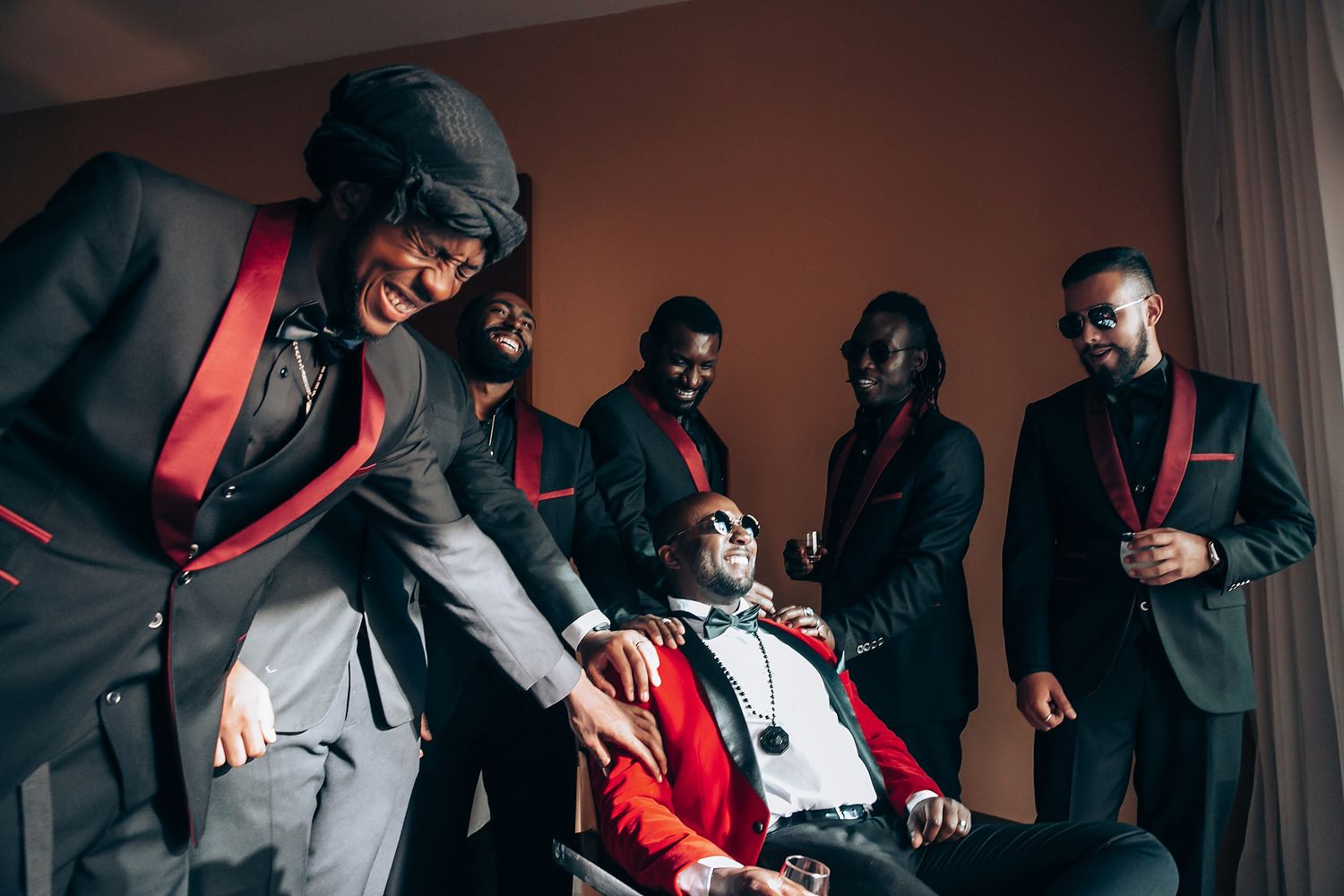 A group in formal black and red attire poses together in a dimly lit room during a stylish photoshoot.