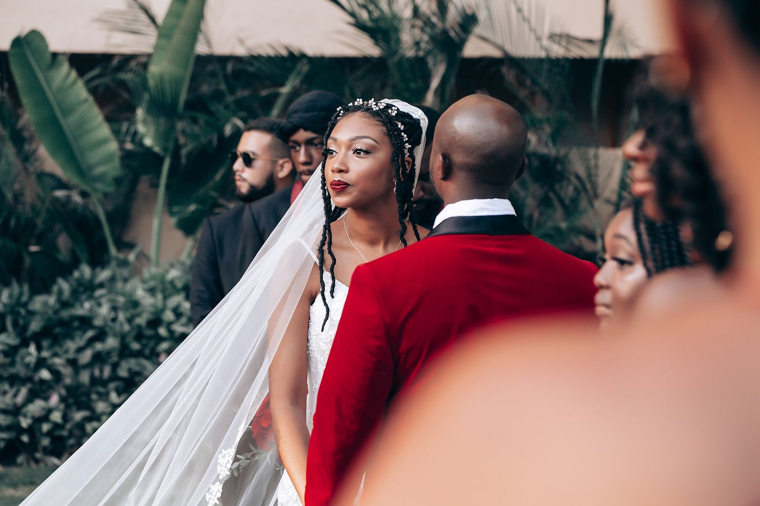 Bride in flowing white veil stands with groom in striking red jacket during outdoor wedding ceremony.