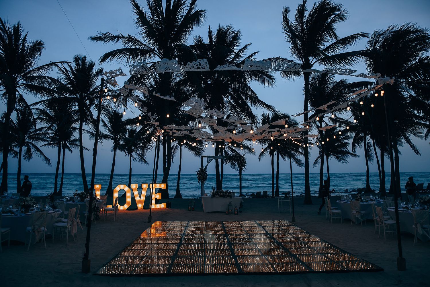 Illuminated LOVE sign glows against silhouetted palm trees at dusk on tropical beach.