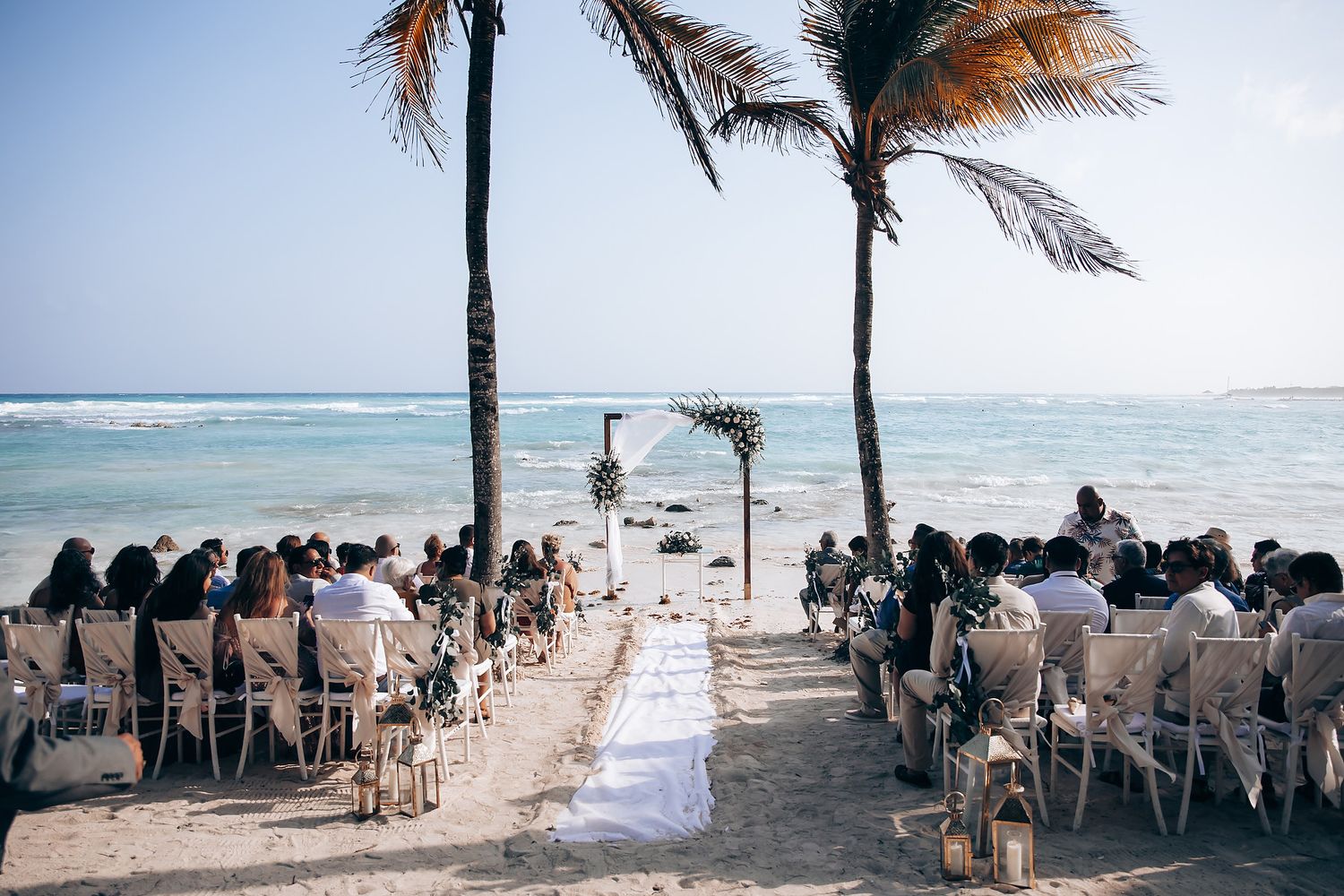 Beachside wedding ceremony setup with white chairs and palm trees swaying in the breeze along a tropical shoreline.