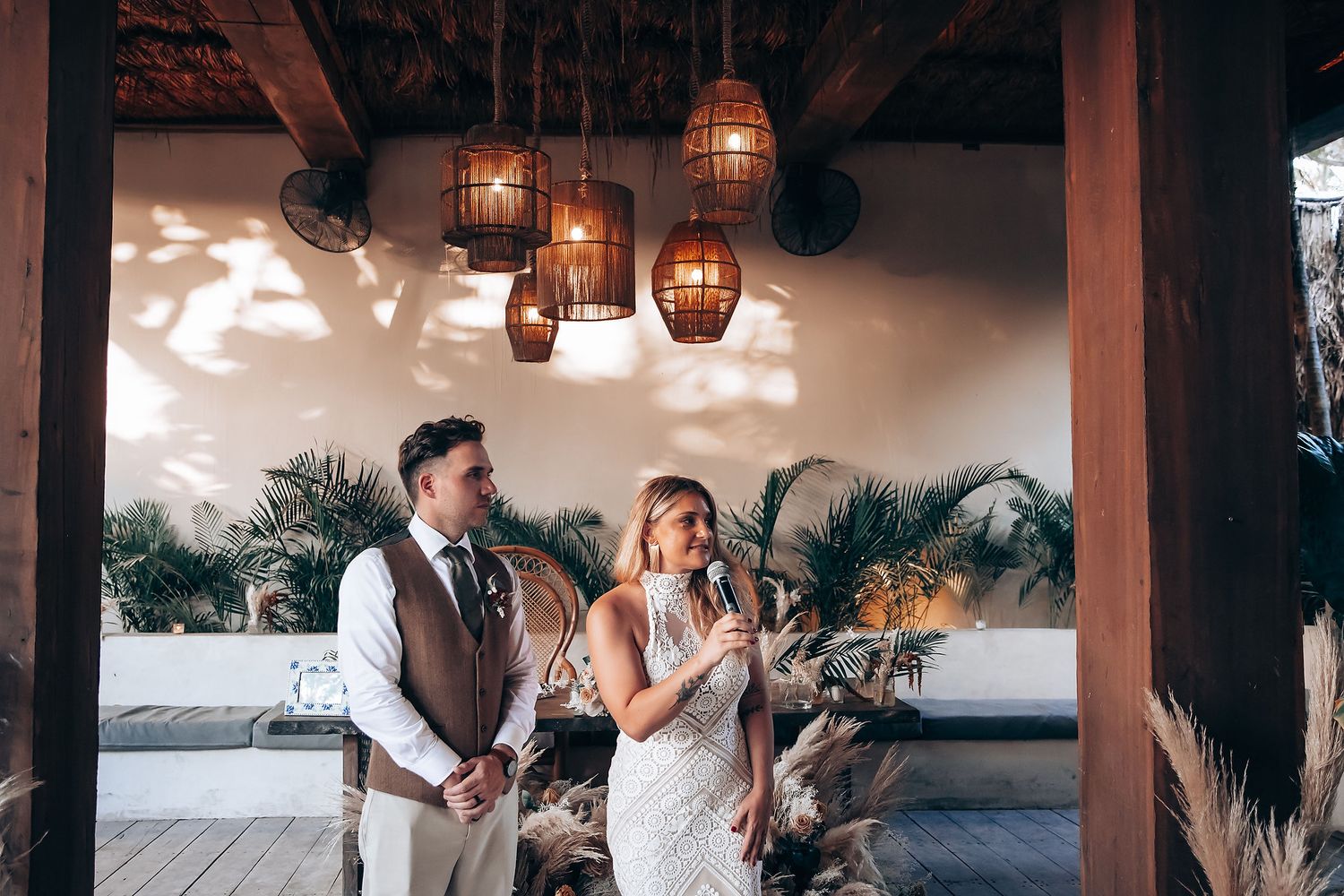 Outdoor wedding reception with rustic wooden tables and hanging lanterns surrounded by tropical foliage.