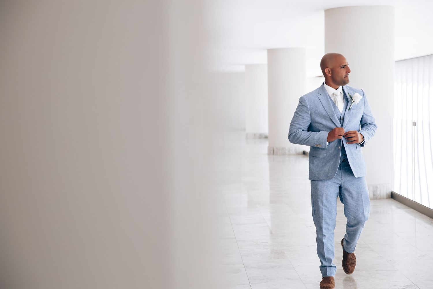 A person wearing a light blue suit walks down a bright white hallway with modern architectural details.