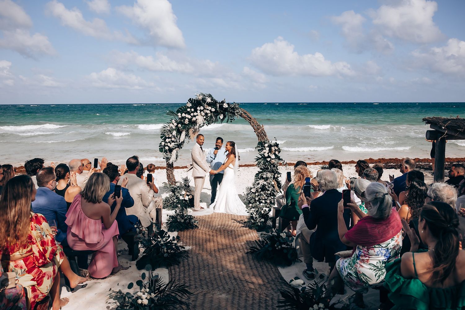 Beachside wedding celebration with decorated arch and guests seated on a sunny day.