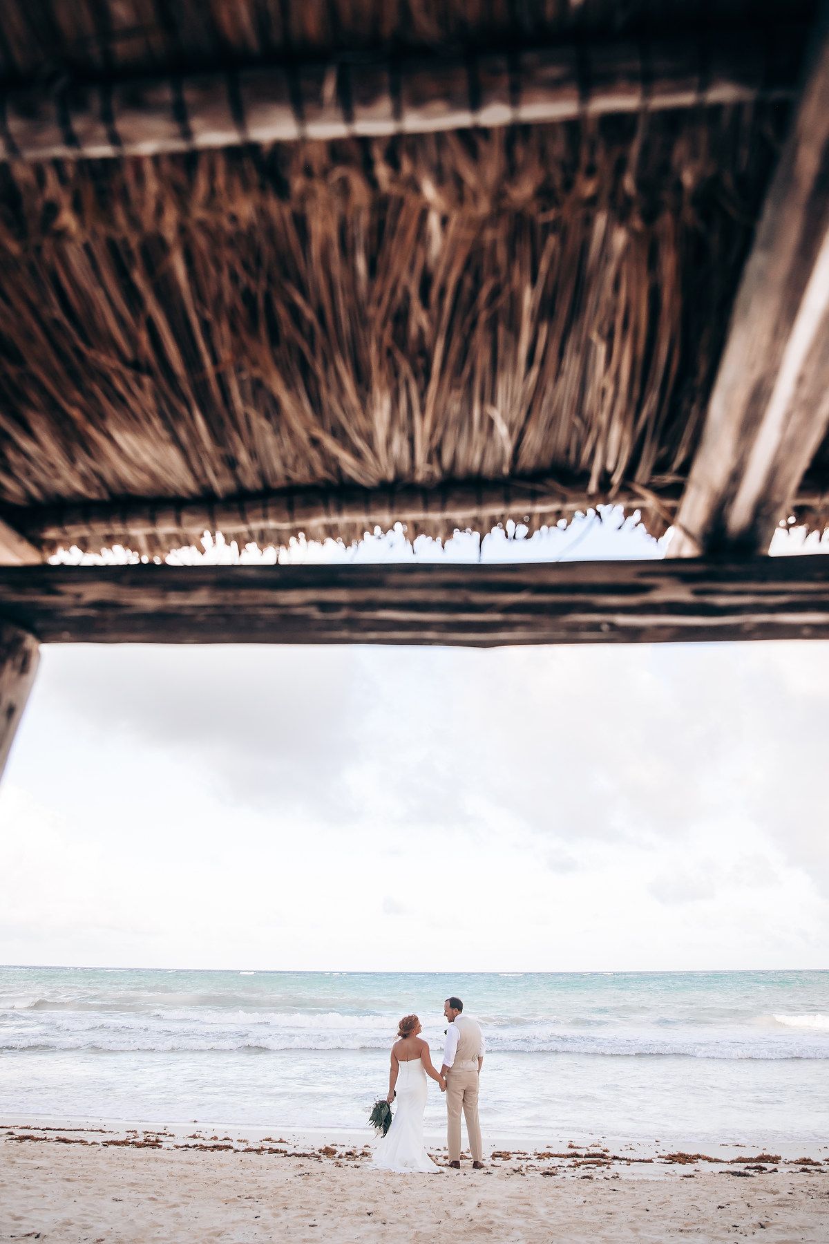 A thatched roof palapa frames a serene beach view with waves rolling onto the shore.