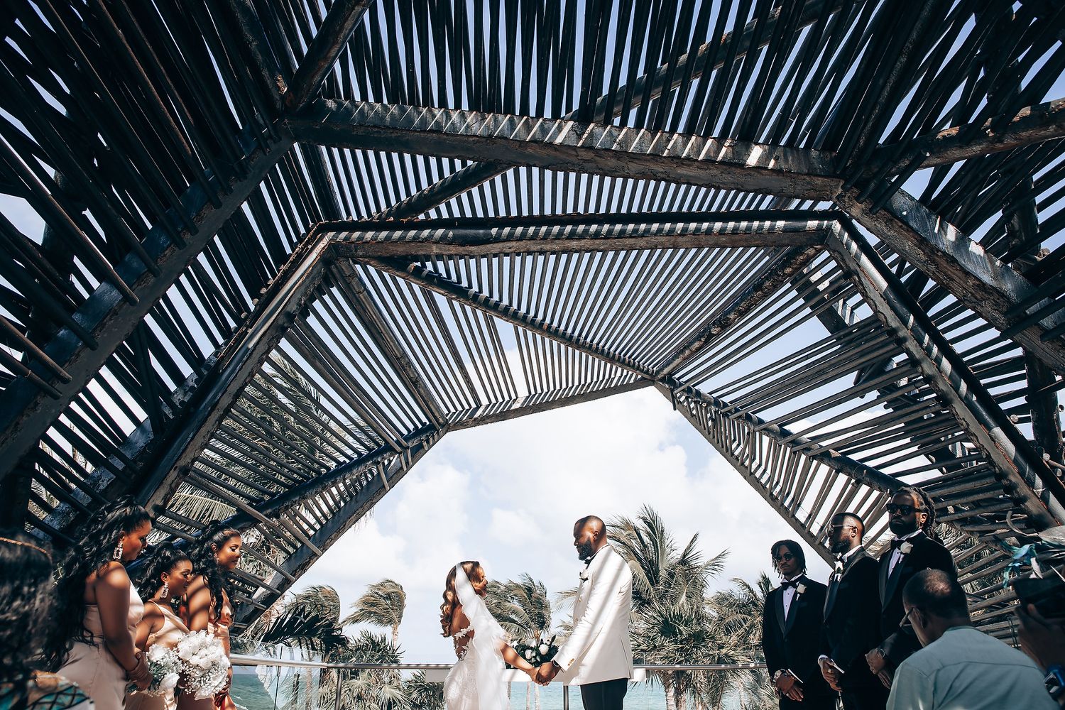 Outdoor wedding ceremony viewed through architectural pergola creating dramatic light patterns.