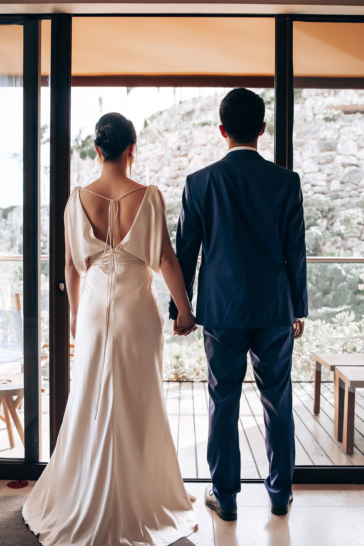 Wedding party members in white dresses and navy suits standing in a modern venue with glass windows.