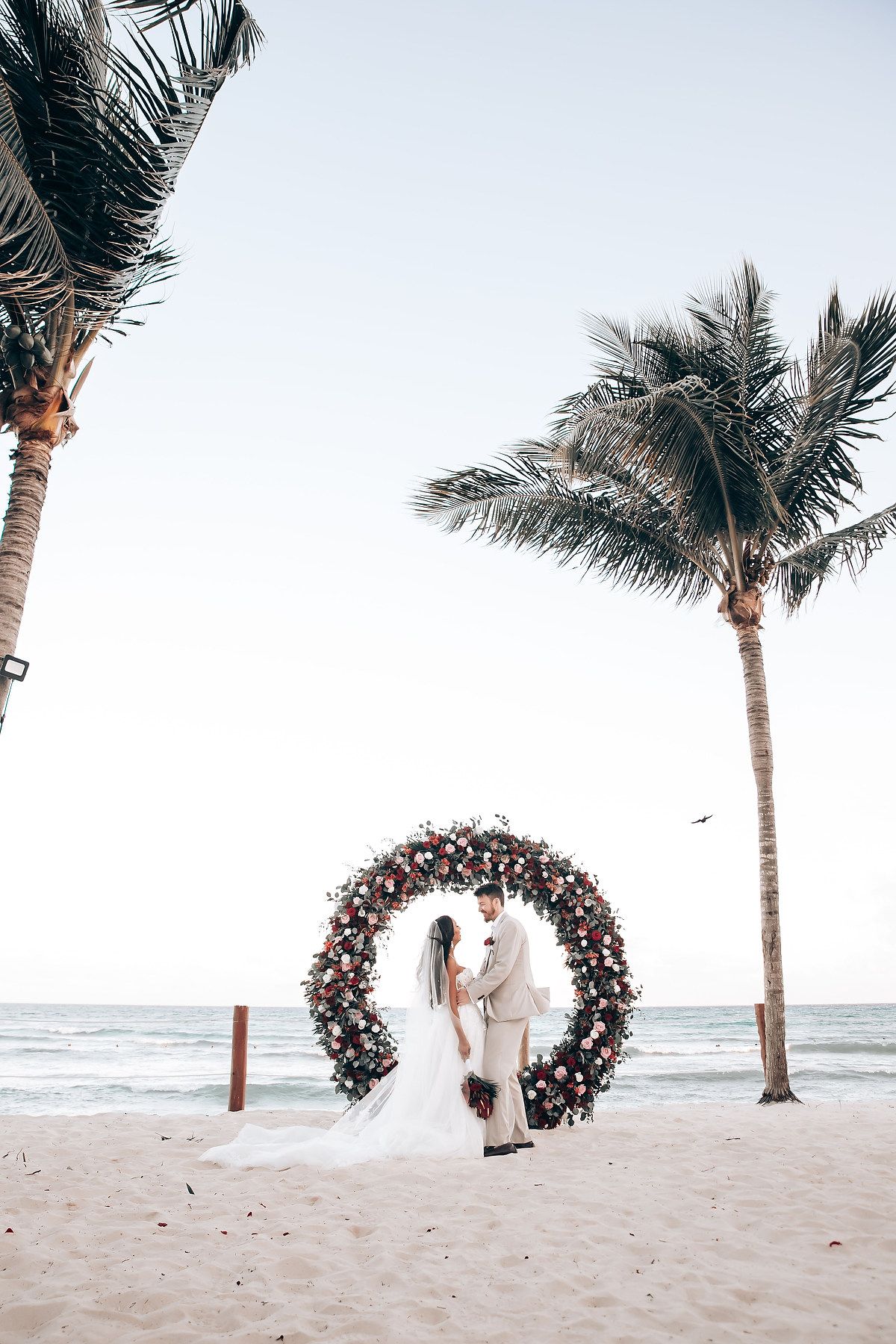 Floral wedding arch on beach with palm trees and ocean view.