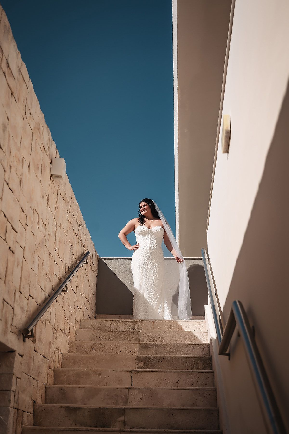 White wedding dress flowing down modern stone steps against a bright blue sky.