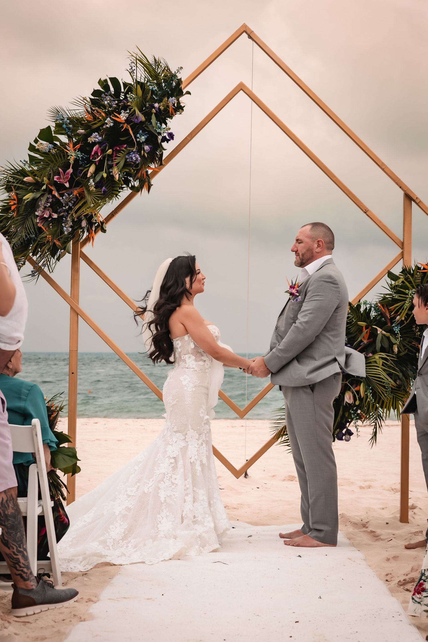 A geometric wooden arch decorated with greenery frames a beachside wedding ceremony.