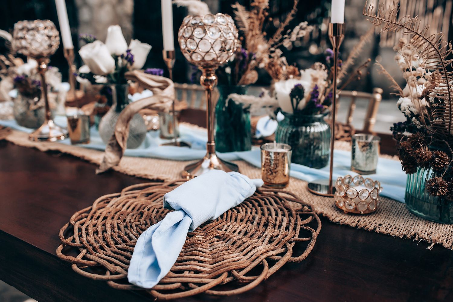 Elegant table setting with woven rattan charger plate, crystal stemware and soft blue decor at a wedding reception.