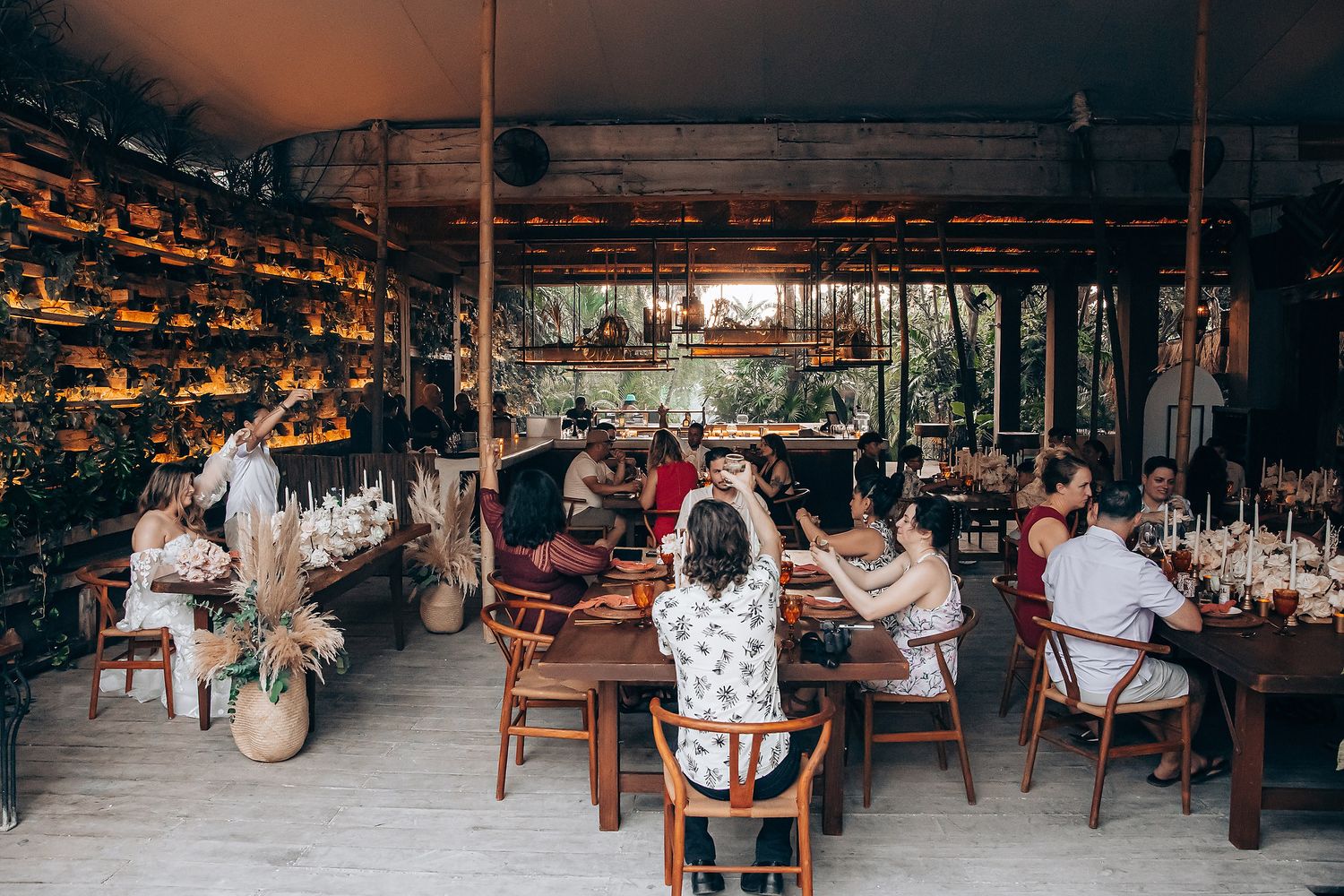 Wedding guests enjoying a lively reception celebration at rustic wooden tables in an open-air venue.