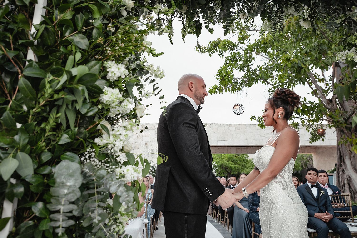 A bride and groom share an intimate moment during their outdoor wedding ceremony under lush green trees.
