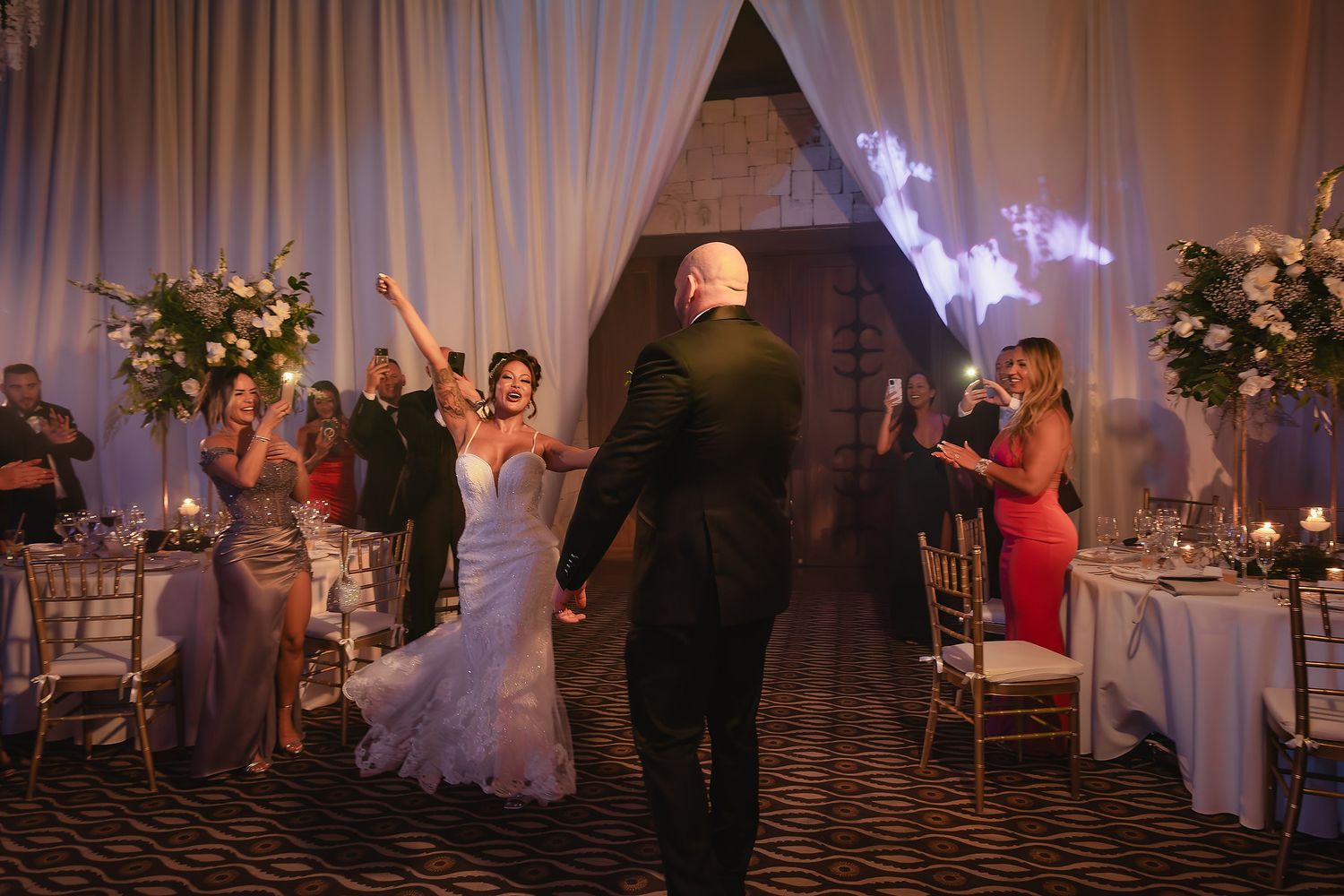 A wedding reception scene with guests dancing in an elegantly decorated ballroom with floral arrangements and white draping.