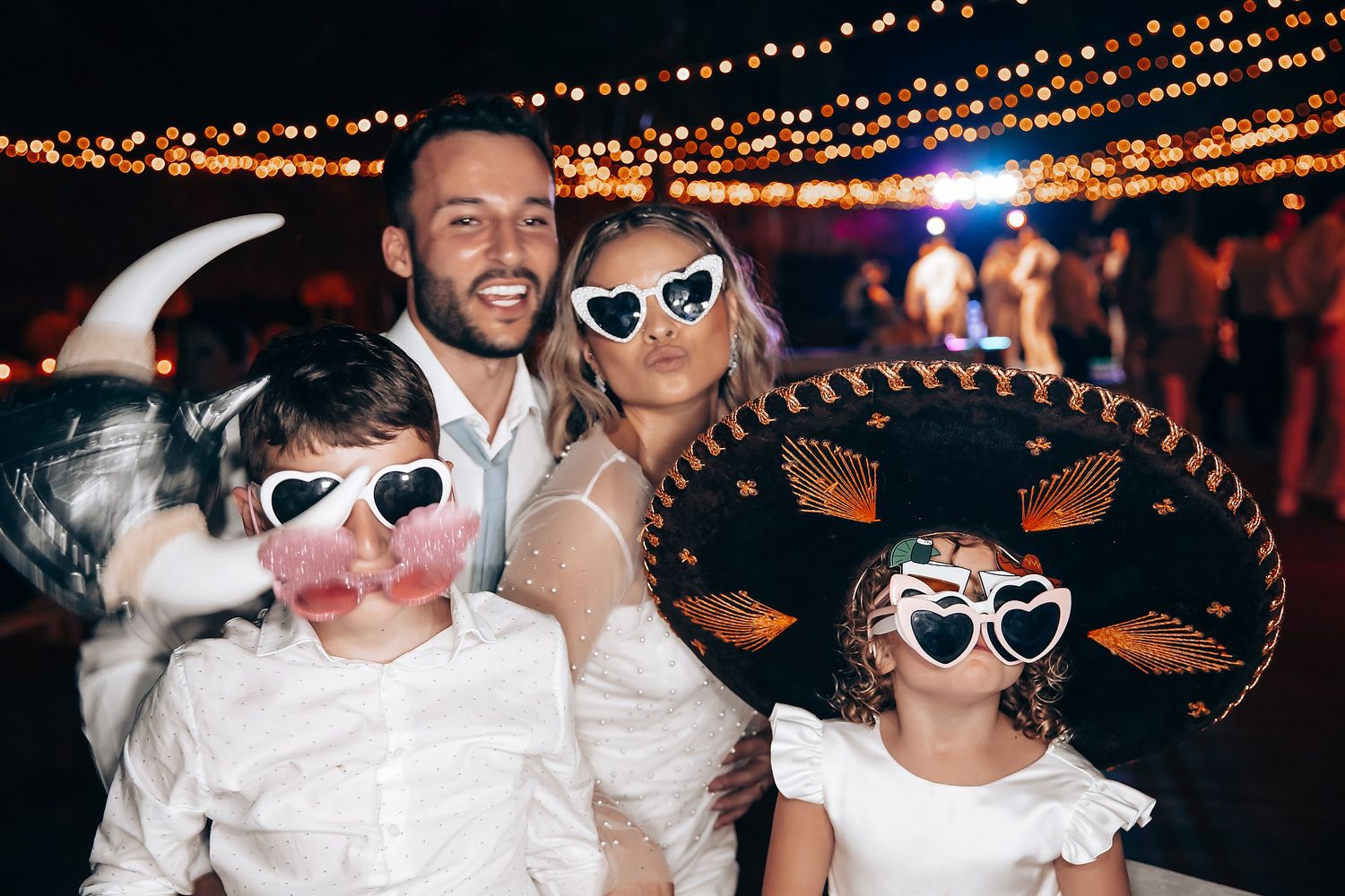 Wedding guests wearing sombreros and sunglasses dancing under string lights at night.