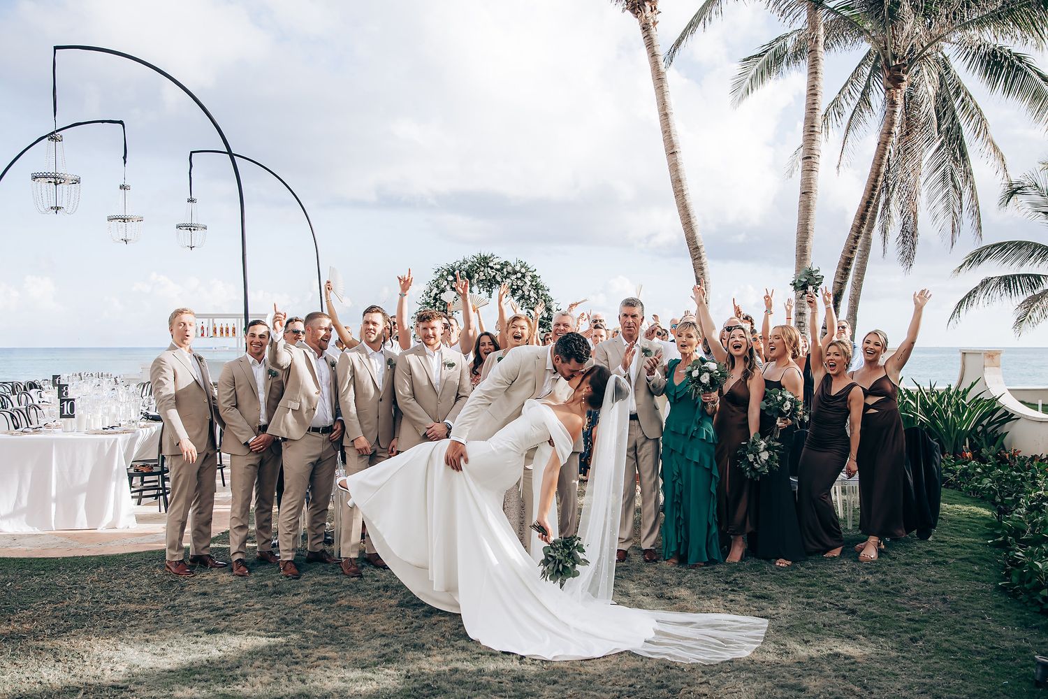 A wedding party celebrates on a tropical beach with palm trees and ocean views during a sunset ceremony.