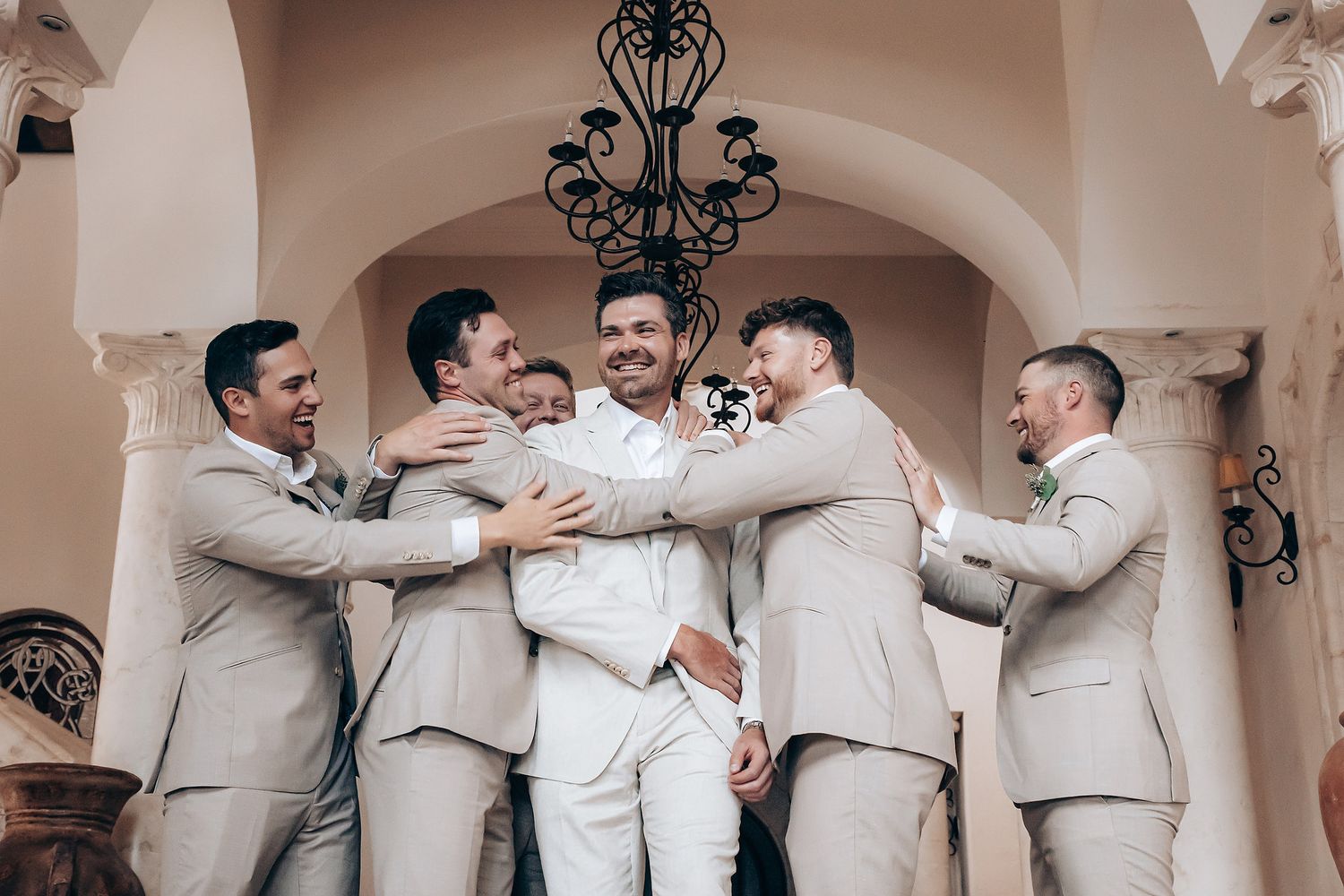Groomsmen in light beige suits gather in a row for candid group photos at an elegant venue.