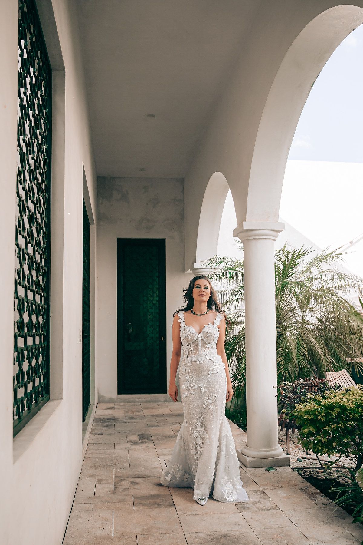 A bride in a white lace gown poses elegantly under a Mediterranean-style archway with white columns and palm trees.