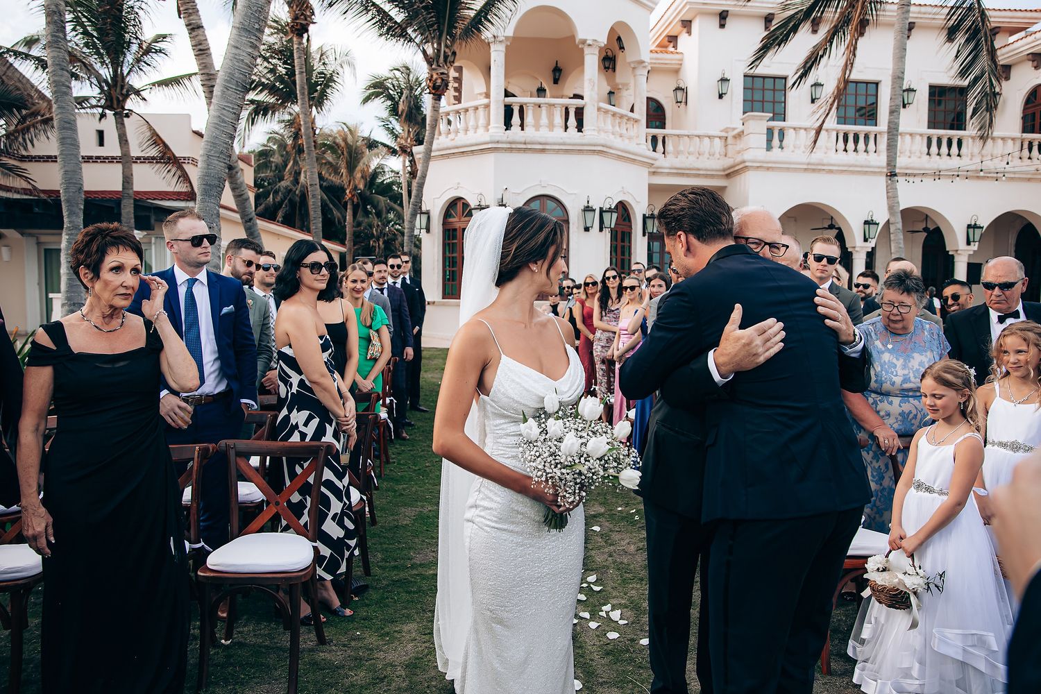Outdoor wedding ceremony takes place on a lawn with palm trees and elegant architecture in the background.