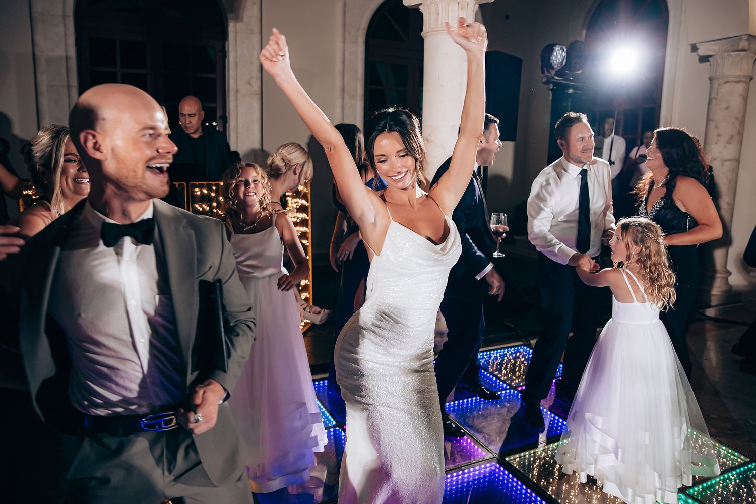Wedding celebration on dance floor with guests dancing under spotlights and decorative lighting.