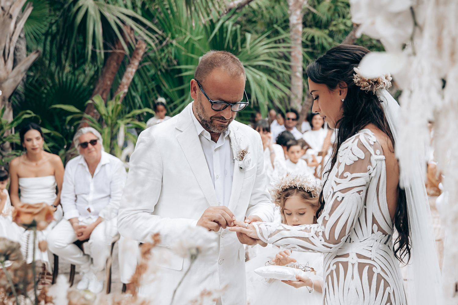Wedding guests dressed in white gather under palm trees and floral decorations for an outdoor celebration.