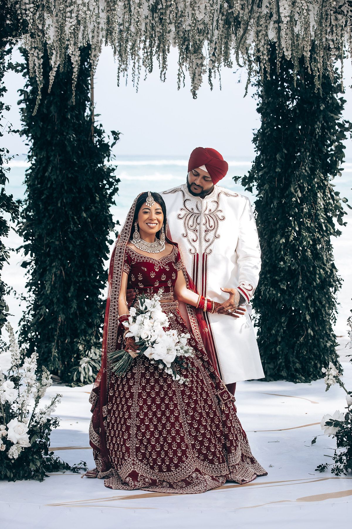 A couple in traditional Indian wedding attire poses under a floral canopy on a beach.