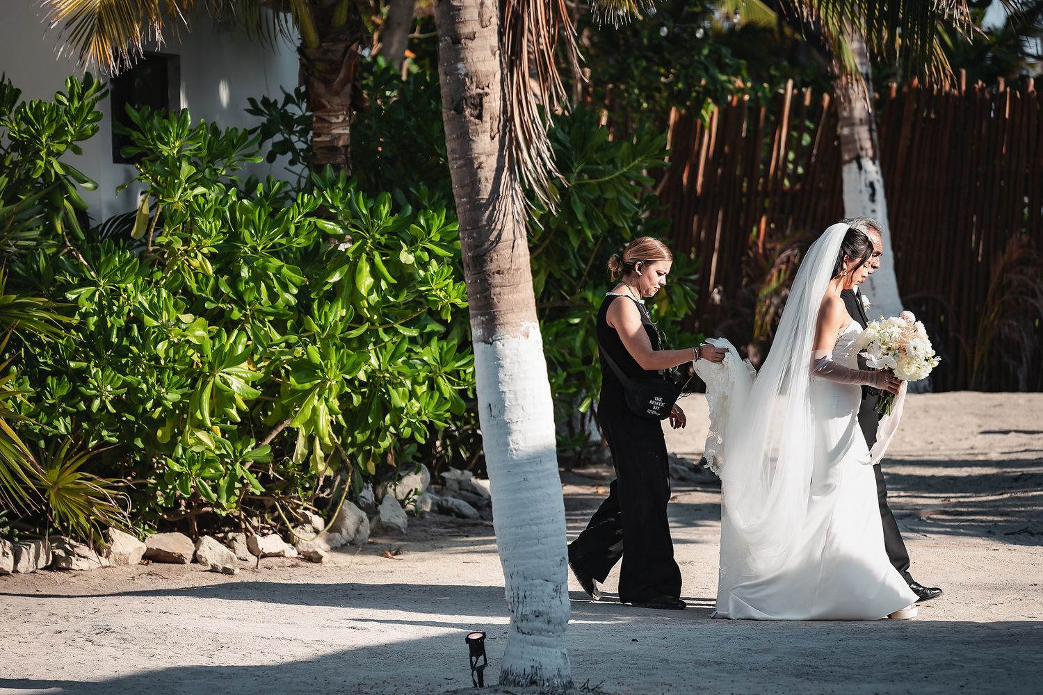 Bride in white gown with long veil walks along sandy beach path lined with palm trees at tropical wedding venue.