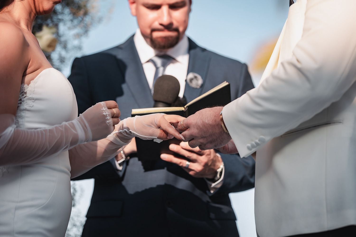 Wedding ceremony with officiant holding Bible, couple exchanging vows outdoors in elegant attire.