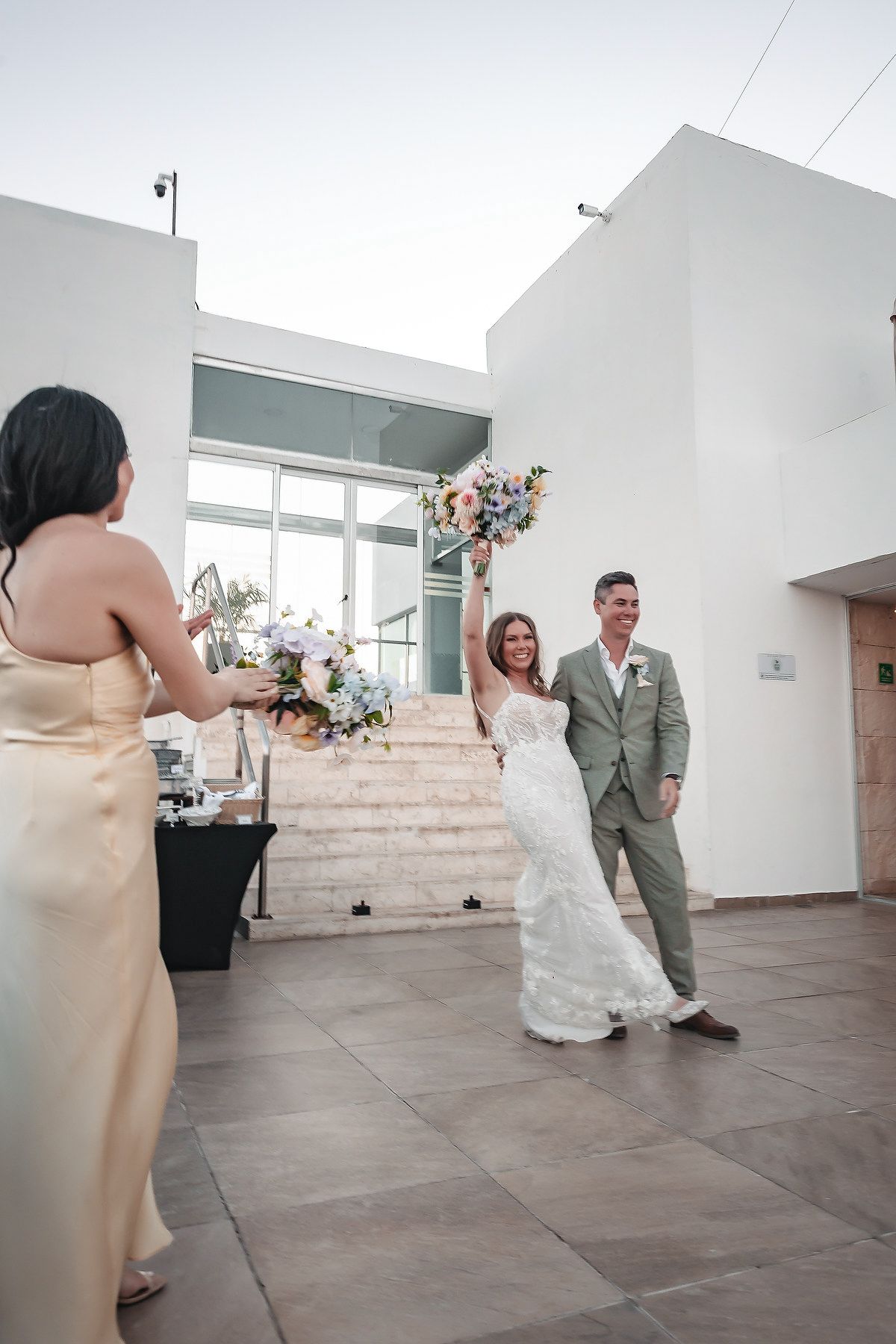 Bride raises bouquet joyfully as groom smiles beside her, greeted by bridesmaid at outdoor wedding venue.