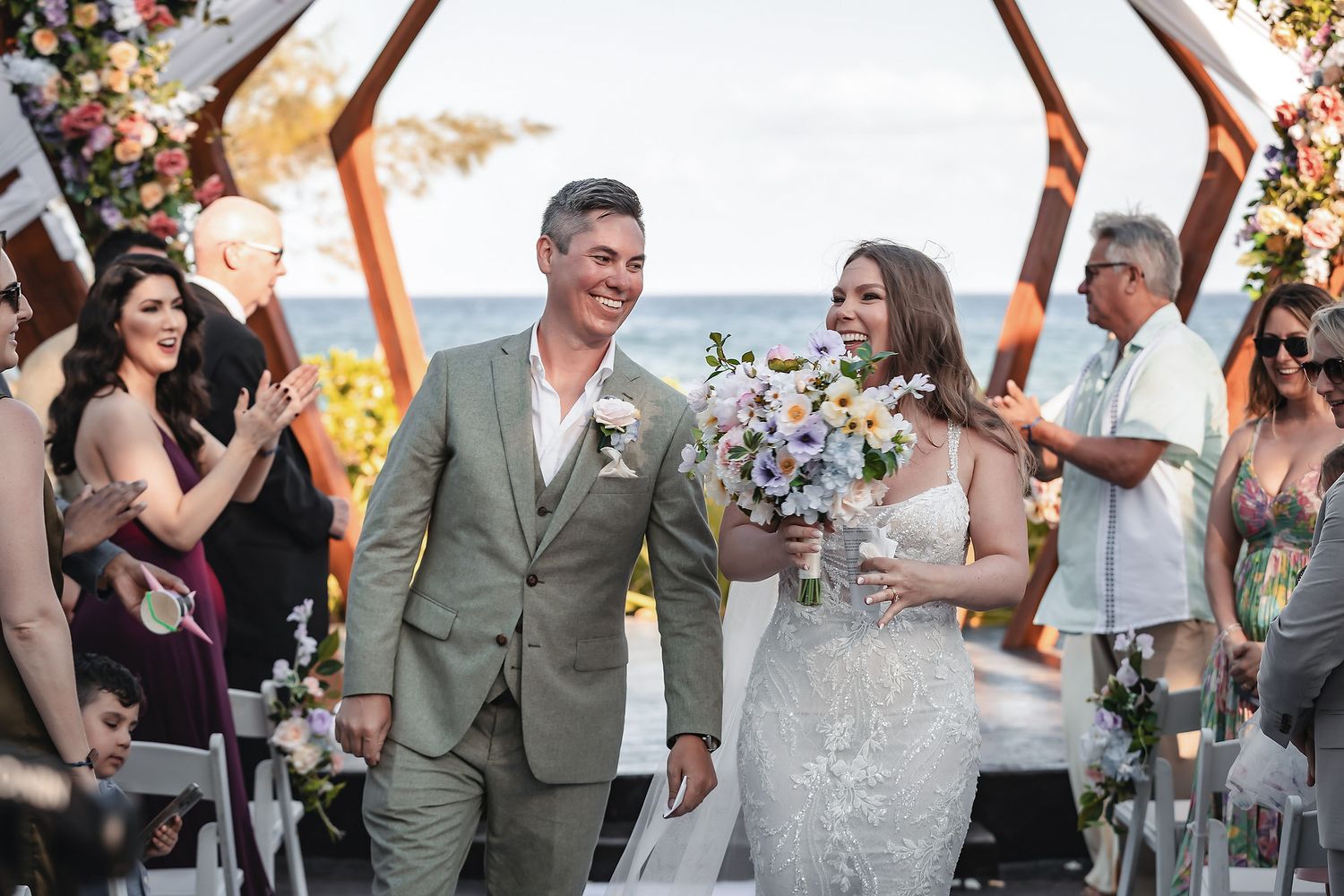 Bride and groom walk down the aisle smiling at outdoor beach wedding ceremony with floral arch.