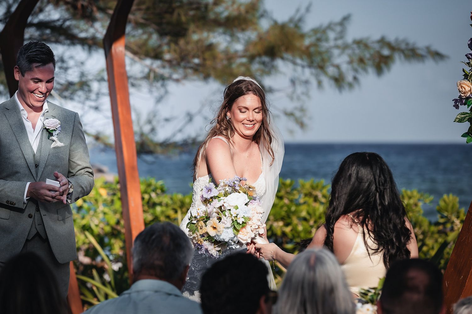 Bride smiling with bouquet at outdoor beach wedding ceremony with groom in gray suit and ocean backdrop.
