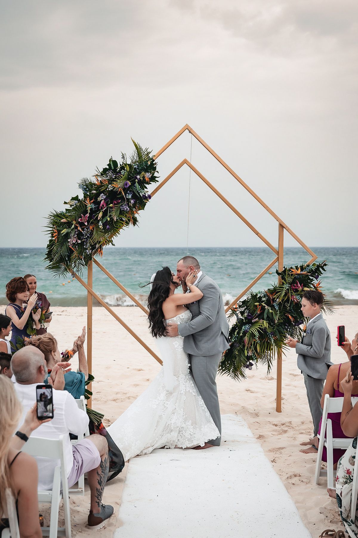 Couple shares first kiss at beach wedding ceremony under geometric gold arch adorned with tropical florals by the ocean.