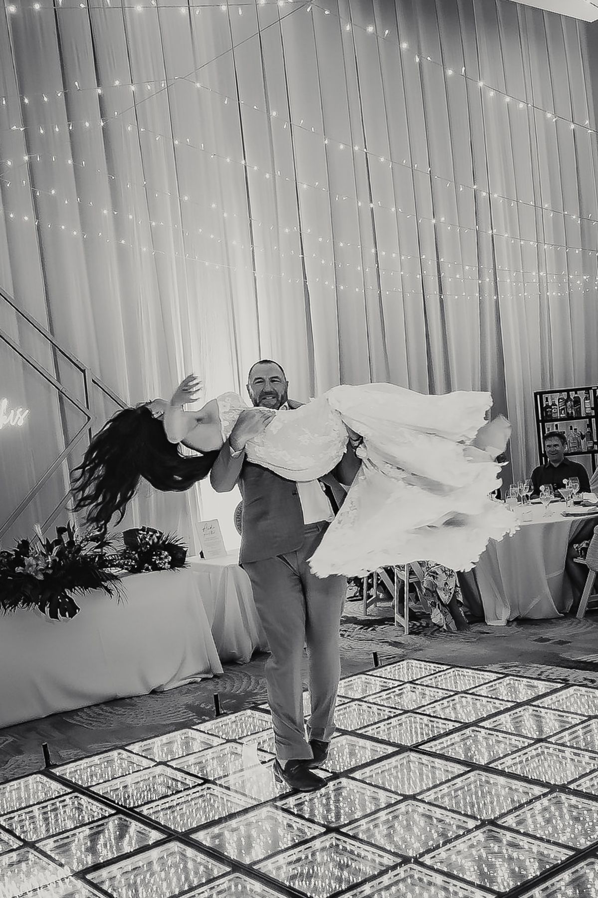 Groom lifts bride in wedding dress on dance floor with fairy lights backdrop in black and white photo.