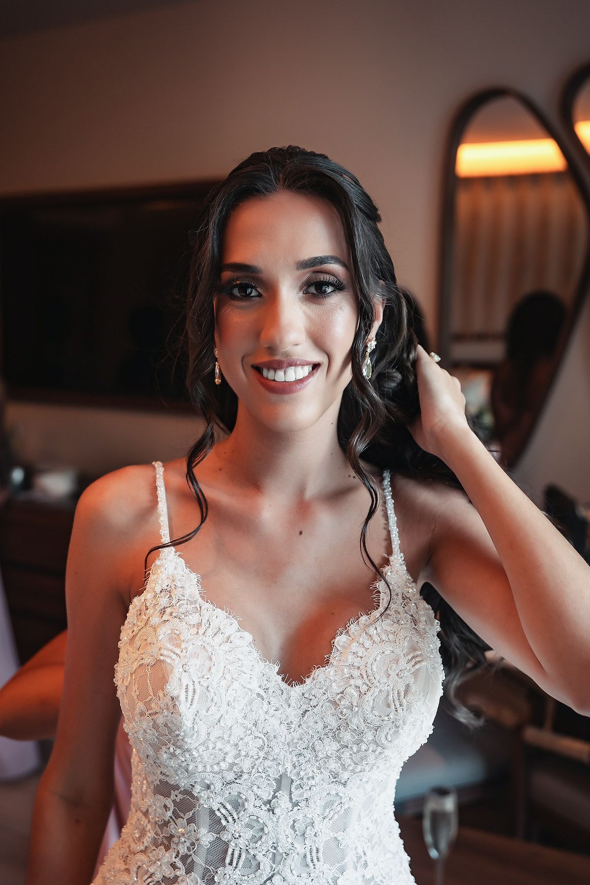Smiling bride in white lace wedding gown adjusting her earring, with elegant makeup and dark wavy hair.