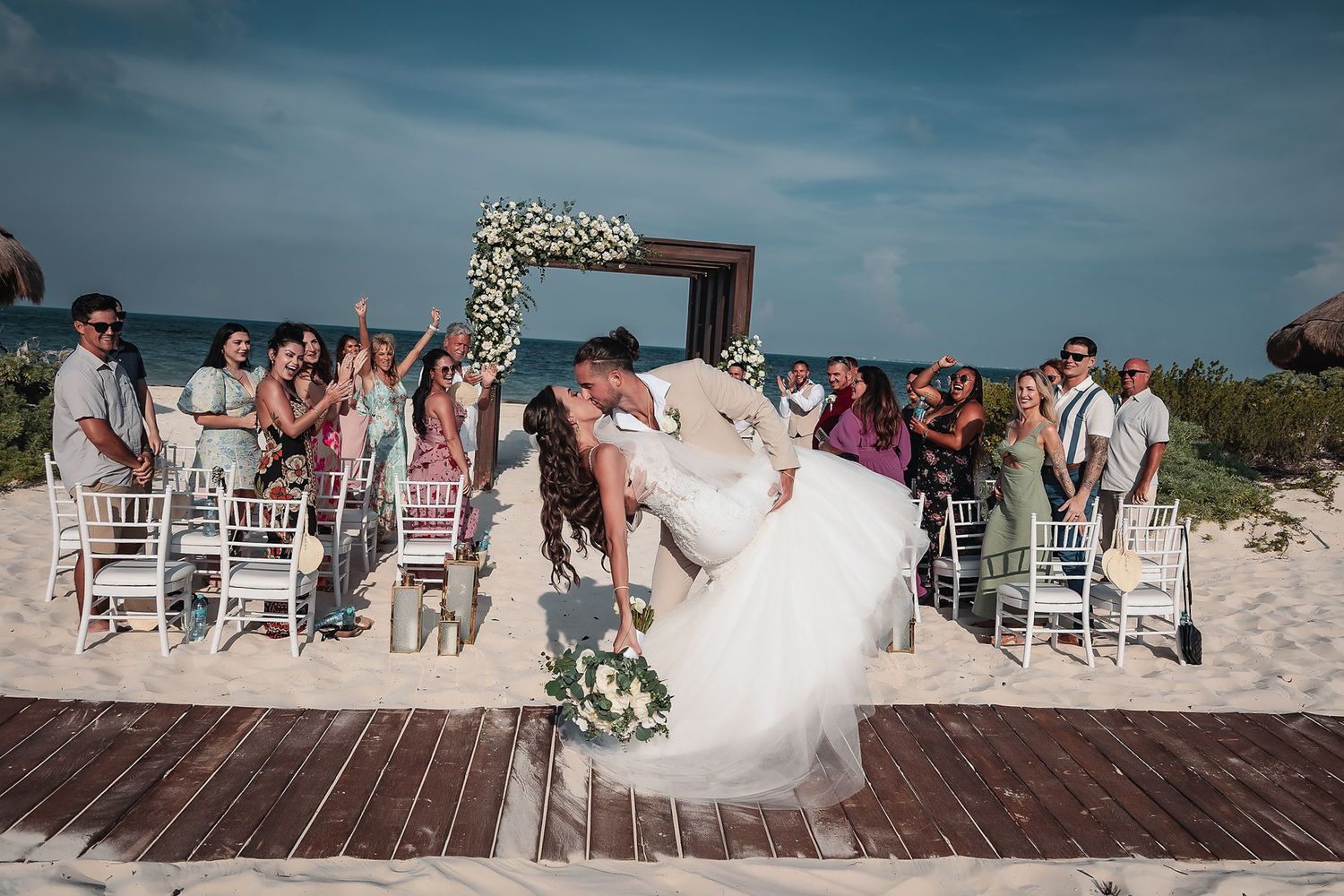 Couple shares romantic kiss at outdoor beach wedding ceremony with guests cheering, floral arch, and ocean backdrop.