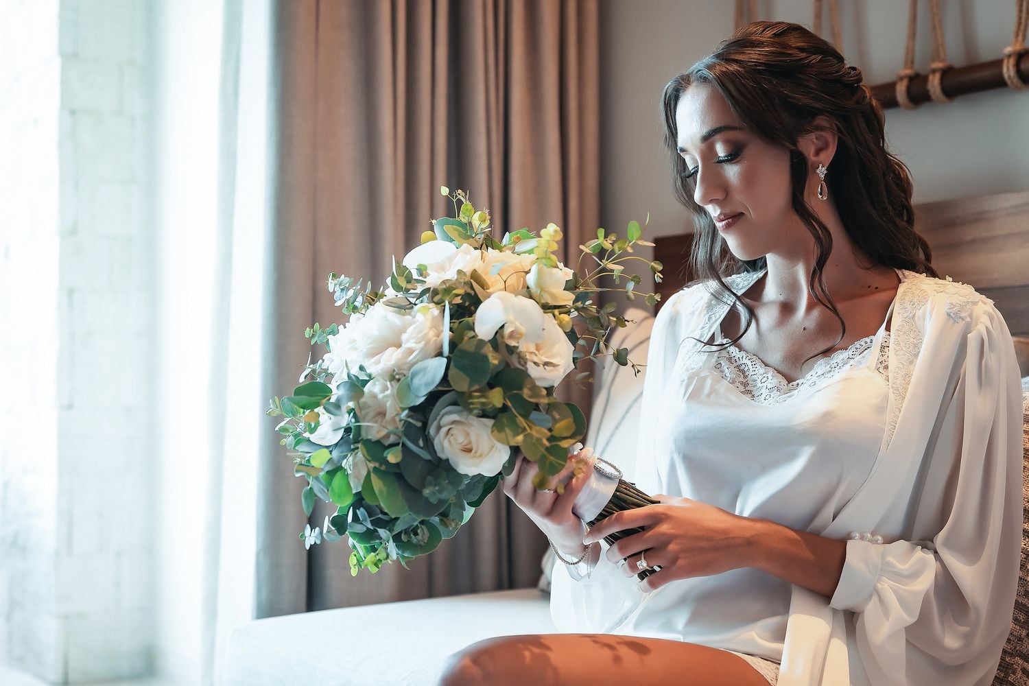 Bride in white robe holding bouquet of white roses and greenery while reading a note by a window.