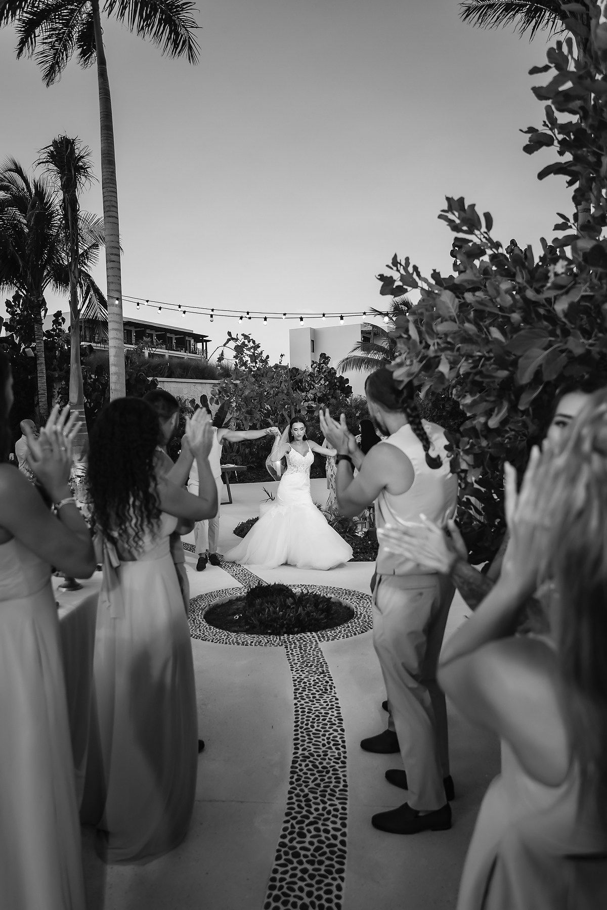 Black and white photo of bride walking down aisle surrounded by clapping wedding guests at outdoor tropical venue.