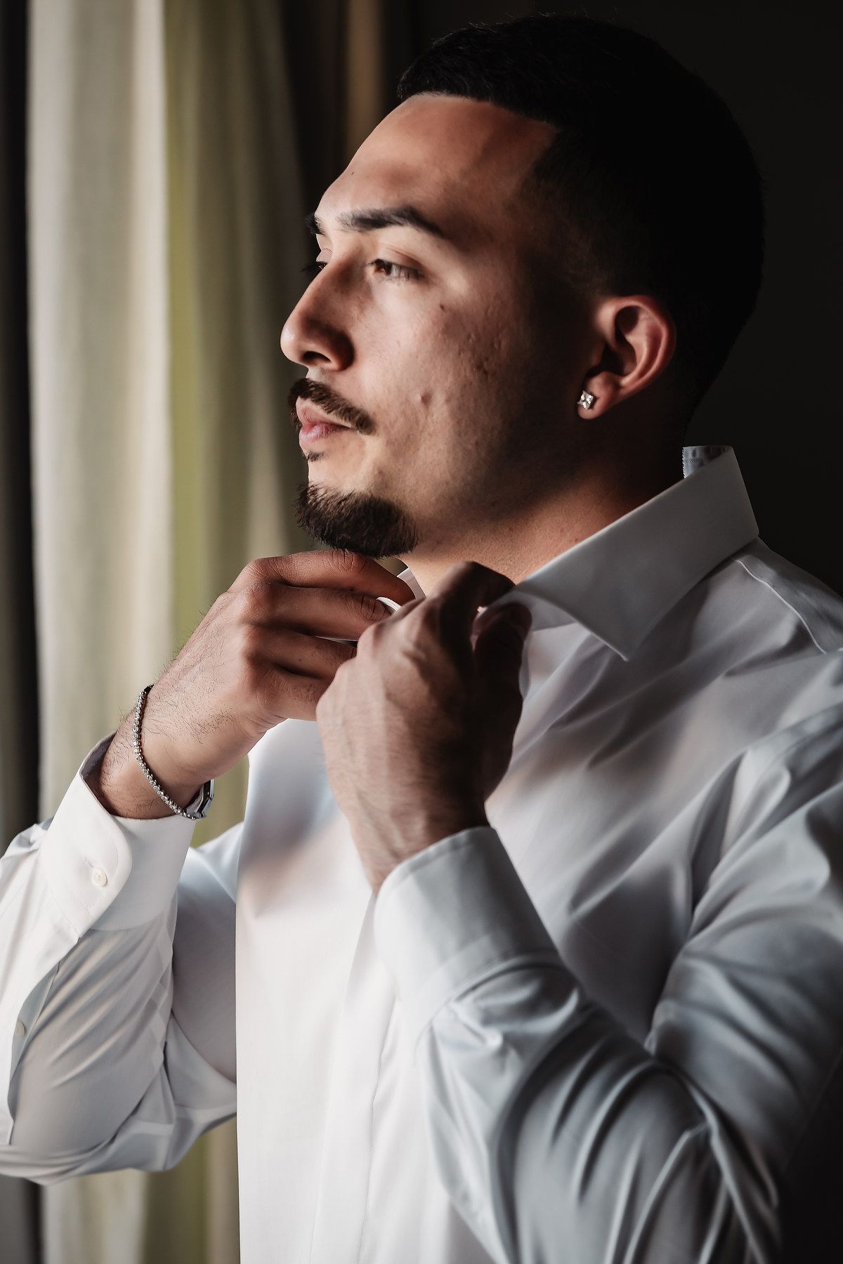 Groom in white dress shirt adjusting collar near window before wedding ceremony.