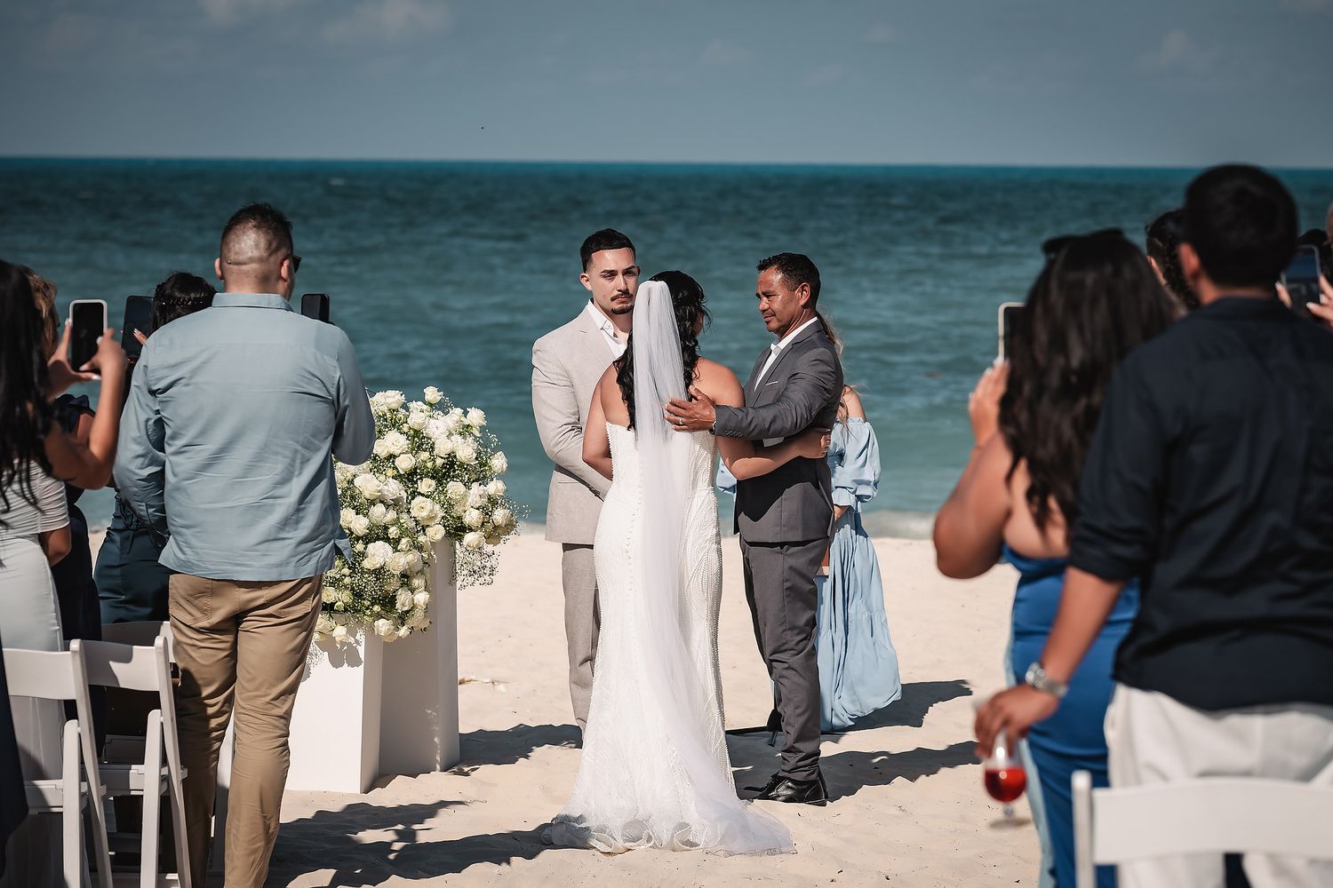 Couple exchanges vows at a romantic beach wedding ceremony with ocean views, white floral arrangements, and guests looking on.