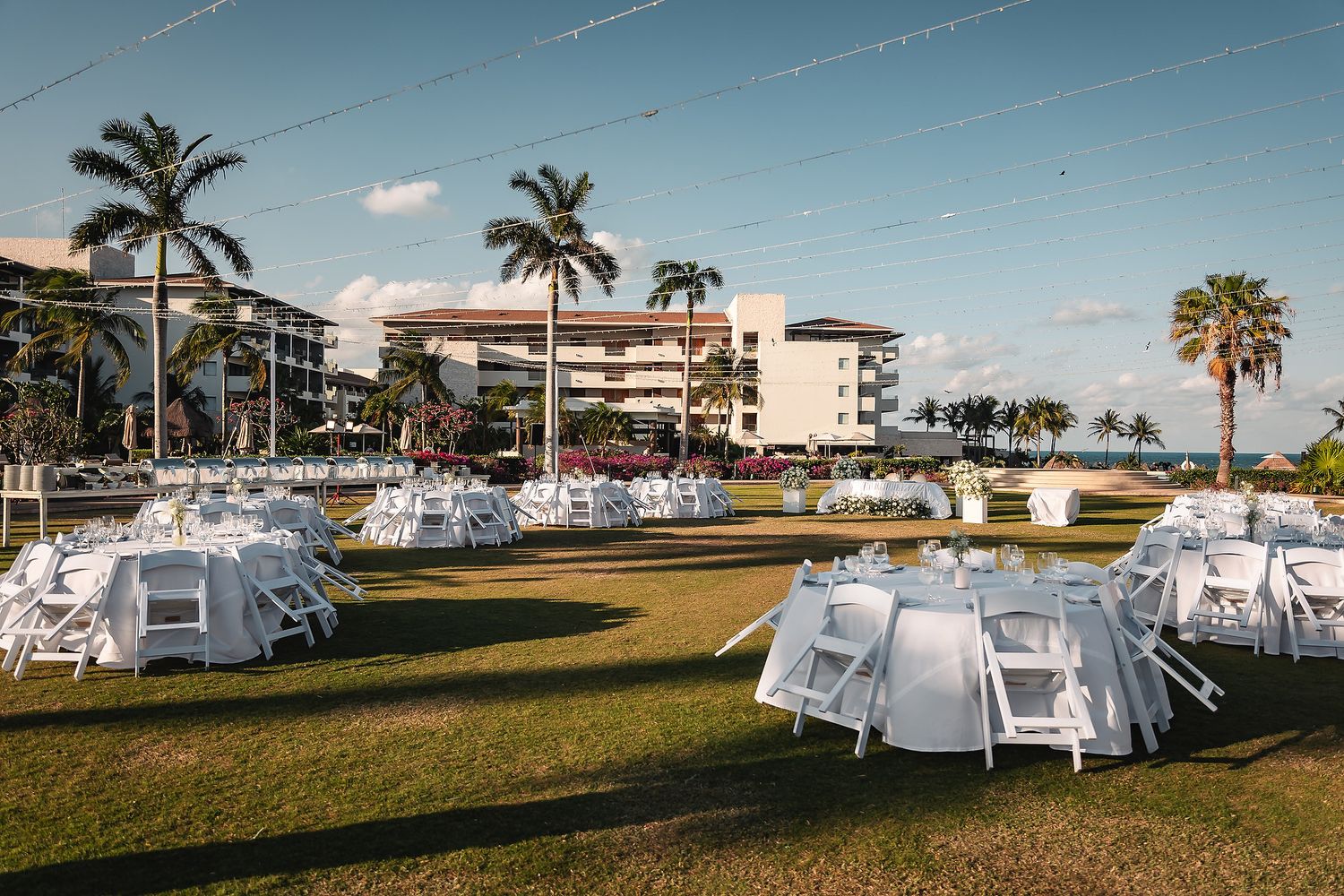 Outdoor wedding reception setup with white chairs and tables on green lawn near a resort with palm trees.