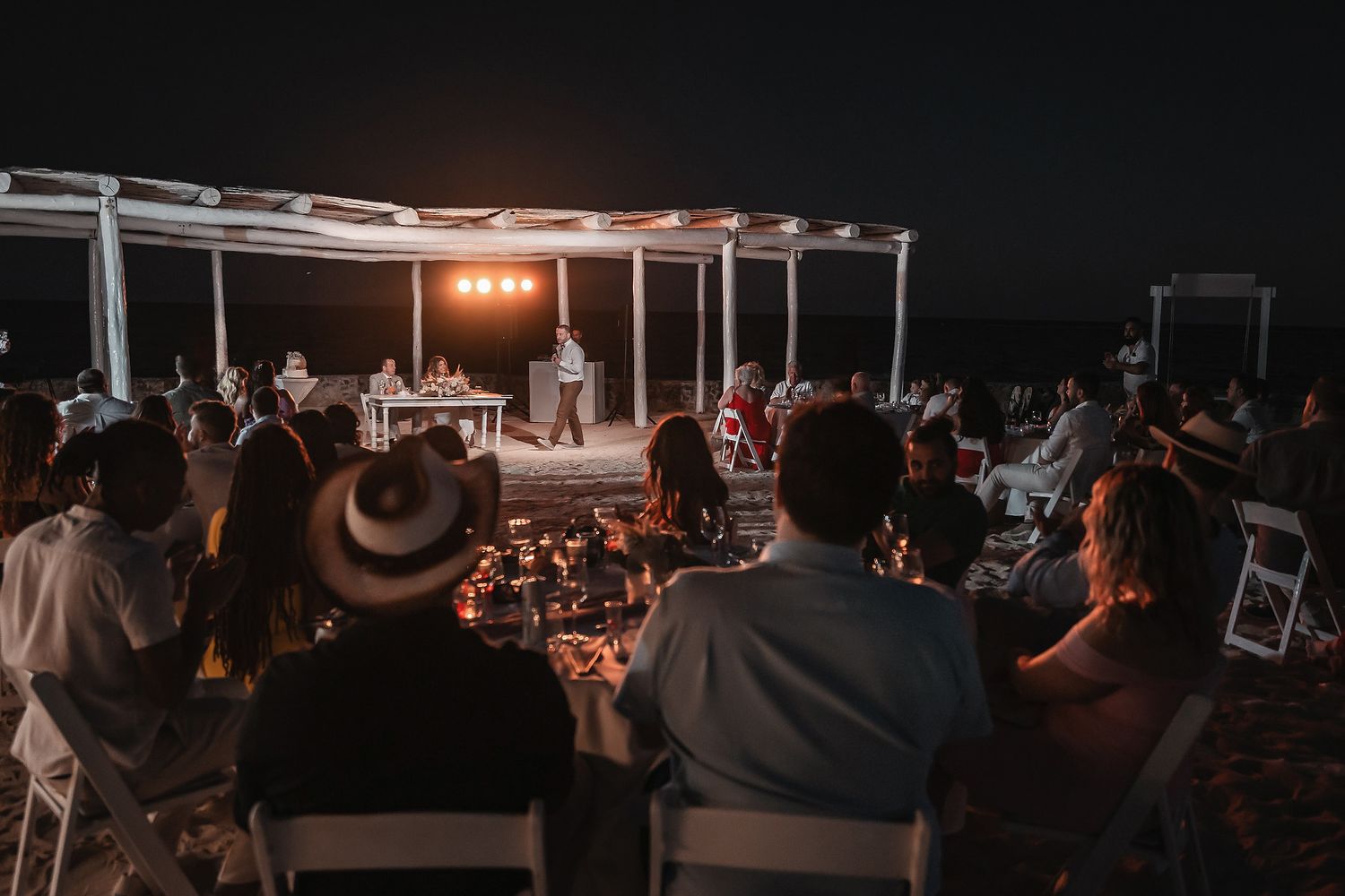 Guests seated at an outdoor beach dinner event at night with a lit stage and pergola in the background.