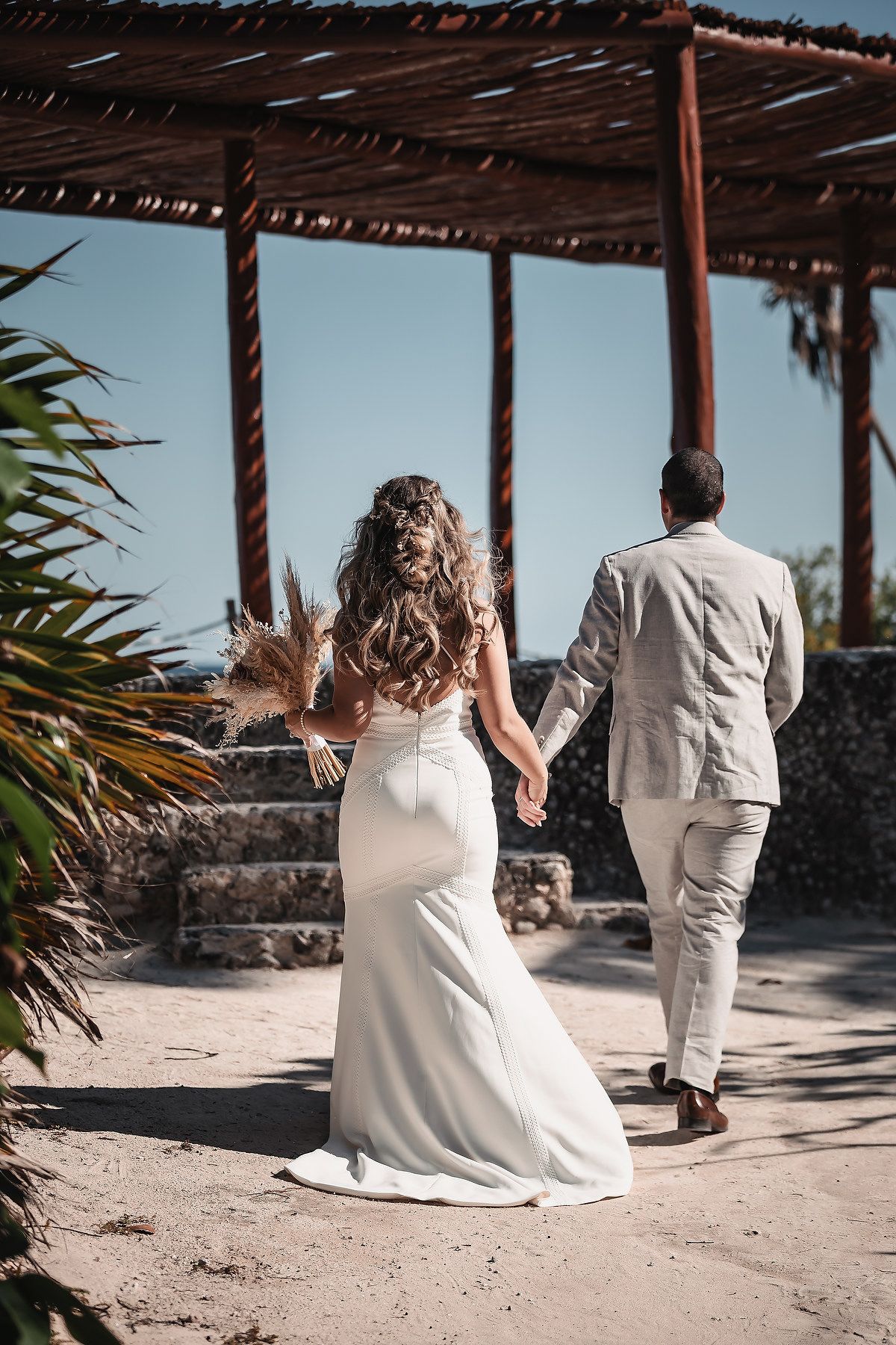 Bride and groom walking hand in hand toward a rustic beach pergola on their wedding day.