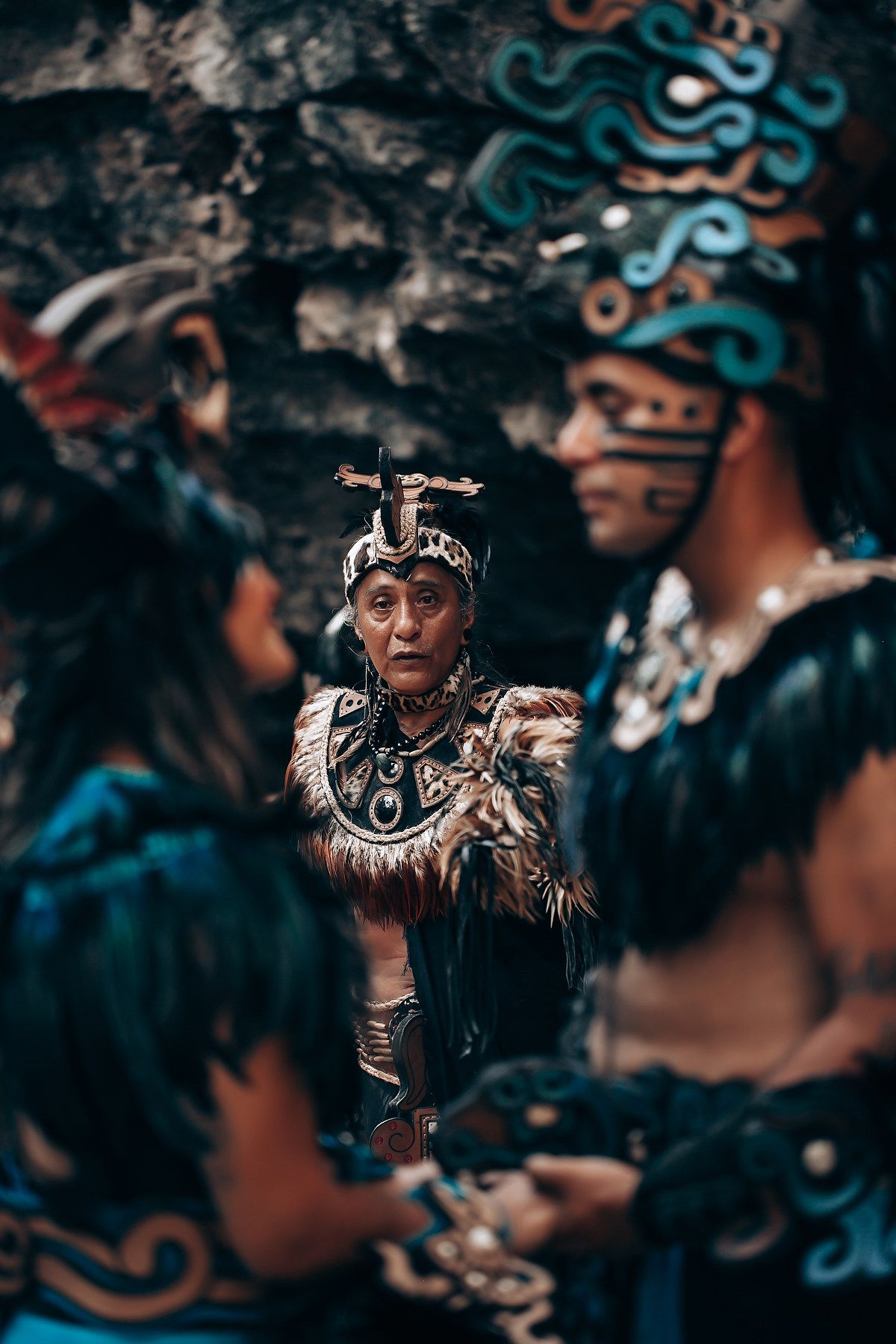 Elder in traditional Mayan ceremonial costume with feathers speaks to performers in ornate Aztec-inspired regalia.