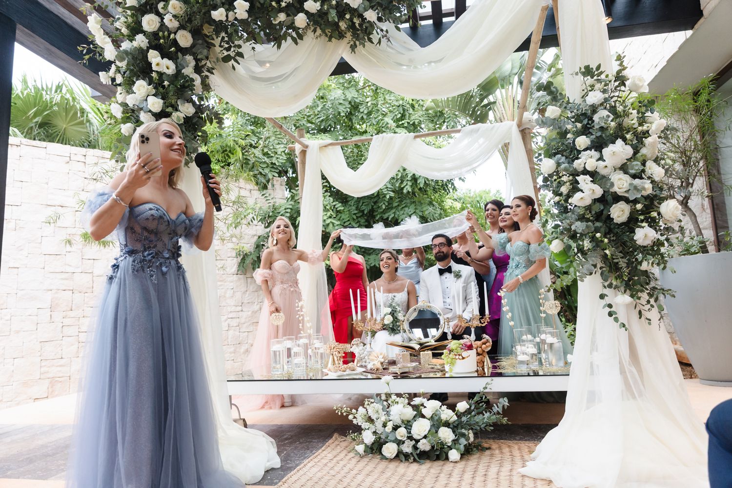 Elegant outdoor wedding ceremony with bridal party under floral arch adorned with white roses and flowing white drapes.