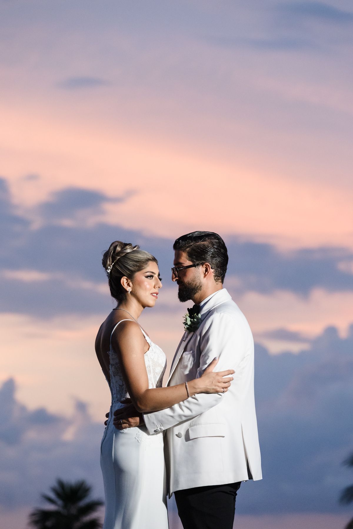 Bride and groom embrace at sunset, wearing white wedding attire against a pink and blue cloudy sky.