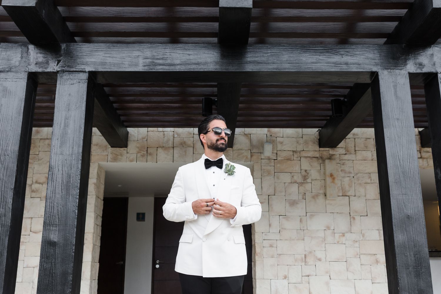 Elegant groom in white tuxedo jacket and bow tie standing outdoors at luxury venue.