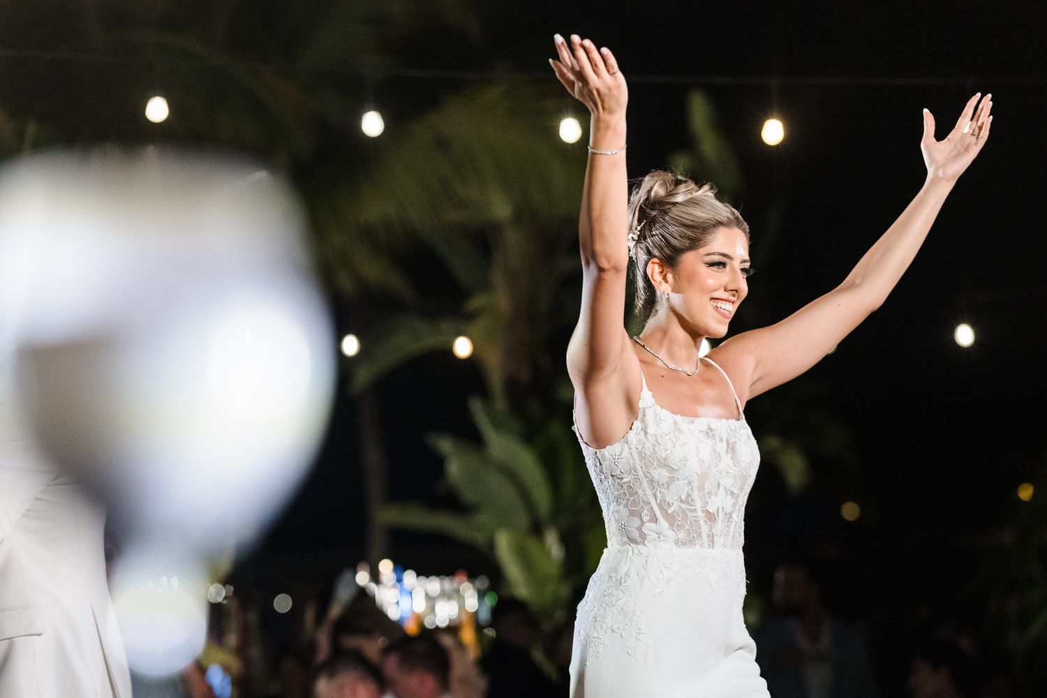 Joyful bride in white gown raises arms celebrating at outdoor evening wedding reception with string lights.