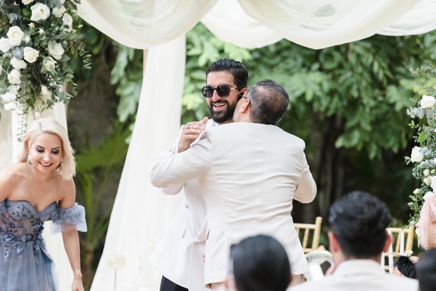 Groom in white suit and sunglasses embracing guest at outdoor wedding ceremony with floral arch.